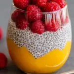 A glass jar filled with creamy chia pudding topped with bright orange mango chunks and red raspberries.
