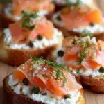A platter of golden baguette slices topped with creamy cottage cheese, pink smoked salmon strips, and fresh green dill.