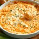 A warm baking dish filled with bubbling healthy buffalo chicken dip topped with melted mozzarella and fresh green onions.