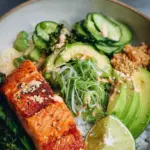 Two bowls filled with crispy salmon cubes, jasmine rice, sliced avocado, cucumber, and edamame.