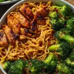 A bowl of spicy garlic chicken with broccoli and noodles topped with green onions and sesame seeds.