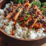 Glazed chicken cubes with steamed broccoli and sesame seeds over white rice in a bowl